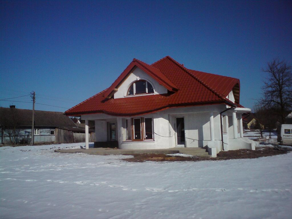 Nowo wybudowany dom jednorodzinny z czerwonym dachem, białą elewacją i charakterystycznym półokrągłym oknem na poddaszu, otoczony śniegiem w słoneczny dzień.